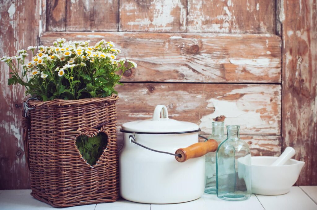 Primitive home decor. Flowers in a wicker basket, glass bottles and vintage milk can on wooden background, cozy home rustic decor, cottage living